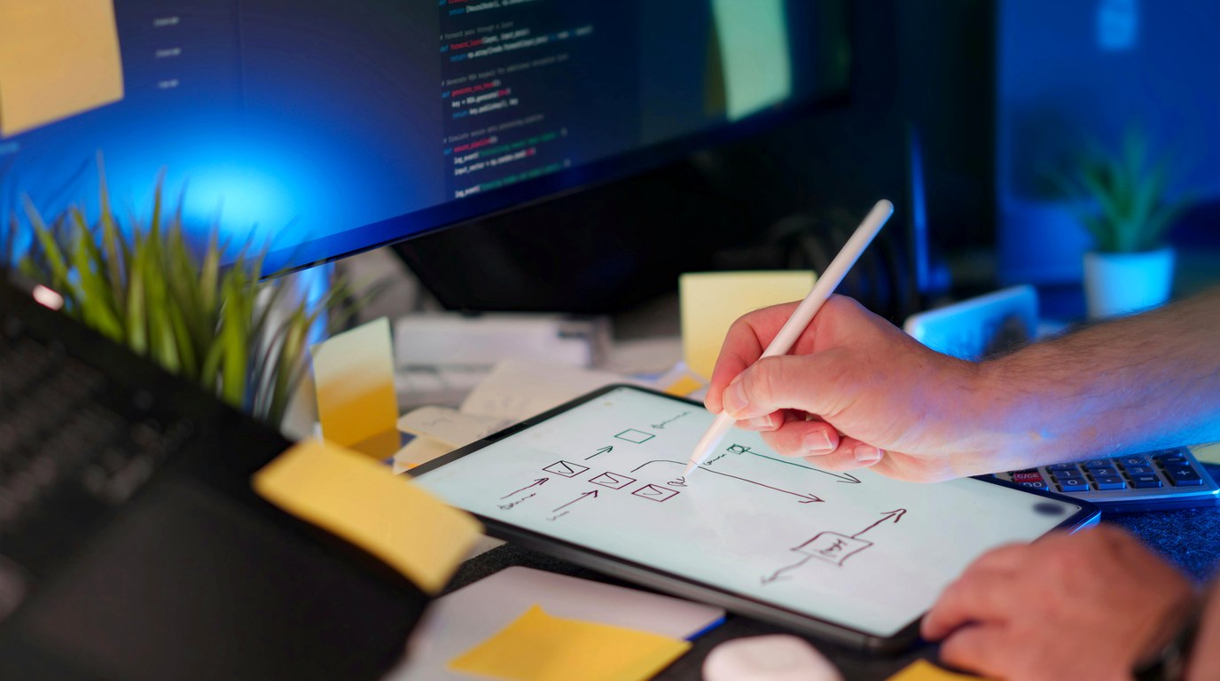 Hand skizziert ein Website-Wireframe auf einem Tablet vor einem Monitor mit Code, Symbolbild für modernes Webdesign