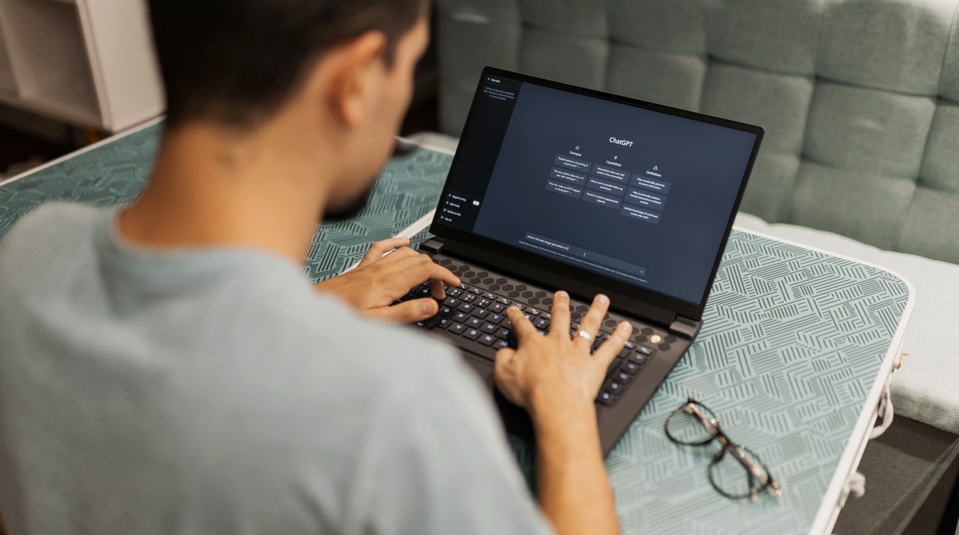 Mann tippt an einem Laptop mit geöffneter ChatGPT-Oberfläche, Brille liegt daneben