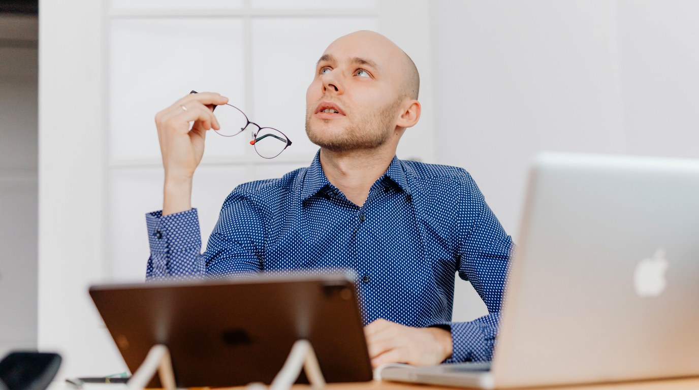 Mann am Schreibtisch mit Laptop hält nachdenklich eine Brille in der Hand und blickt nach oben