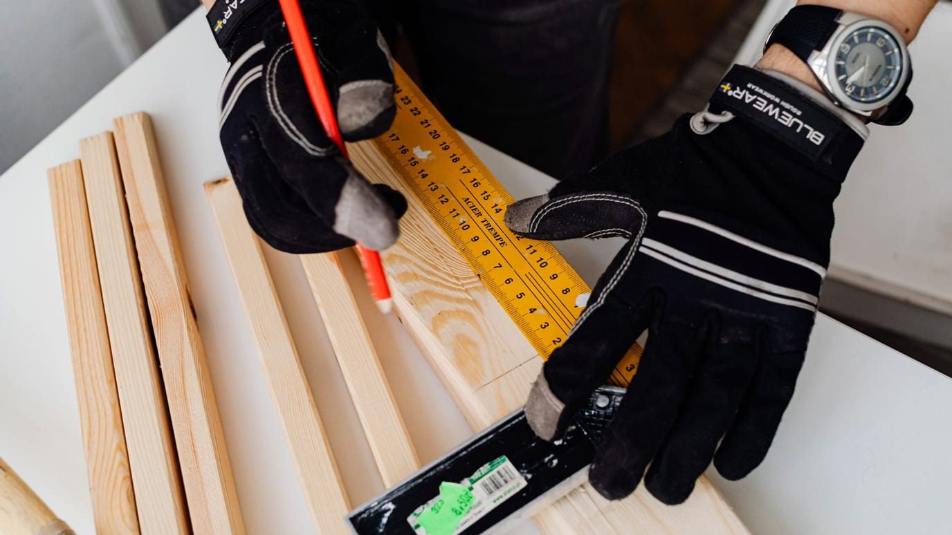 Close-up of gloved hands measuring a wooden batten with a carpenter's square and a red pencil