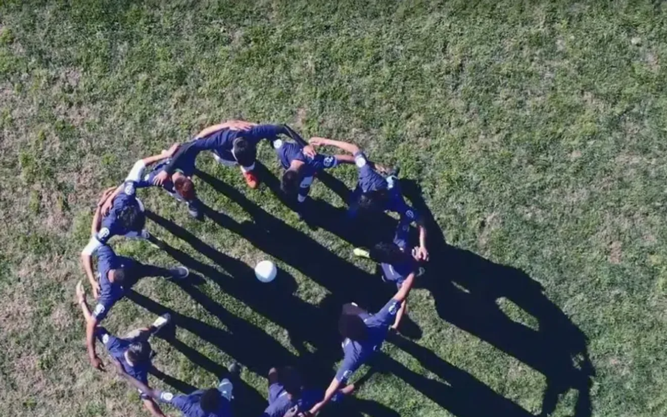 Aerial drone shot of young Manchester City players standing in a circle on a grass pitch, long shadows across the field