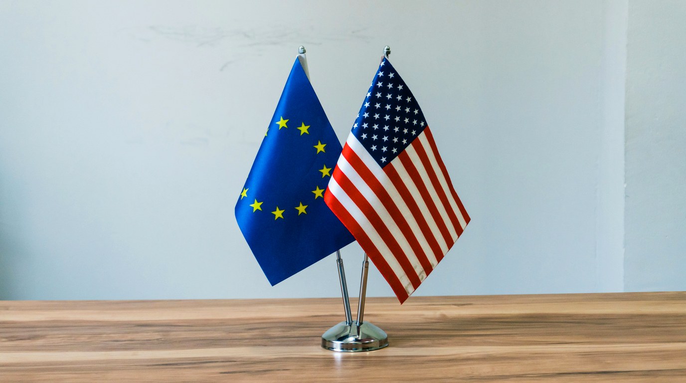 EU-Flagge und US-Flagge auf kleinen Tischstaendern nebeneinander auf einem Holztisch