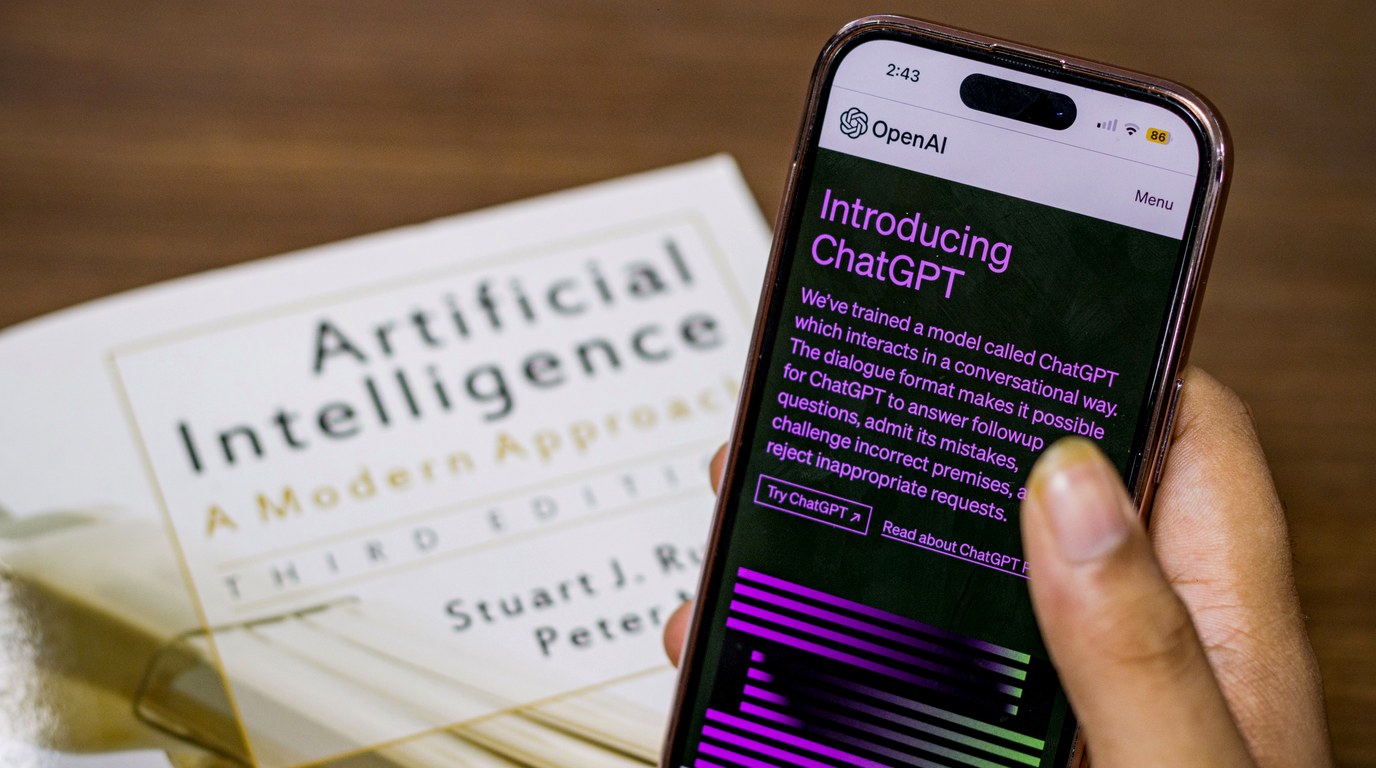 Hand hält Smartphone mit der OpenAI-Ankündigungsseite zu ChatGPT über einem Lehrbuch zur künstlichen Intelligenz