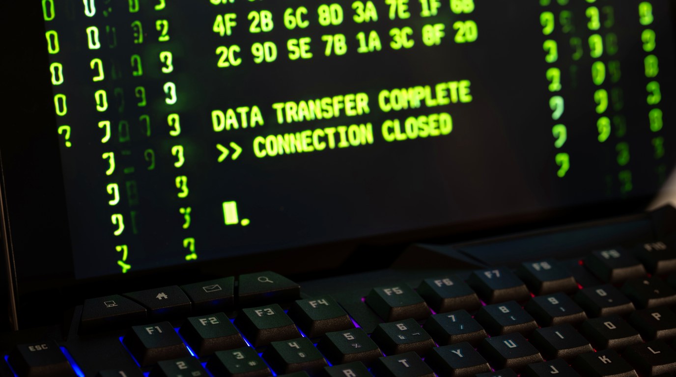 Monitor mit grünem Konsolentext neben einer beleuchteten Tastatur in einem abgedunkelten Raum