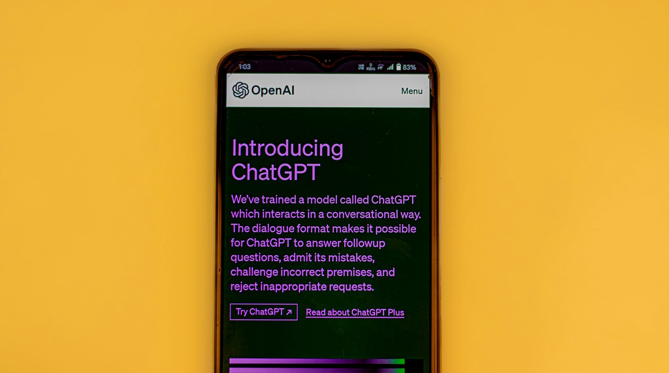 Smartphone auf gelbem Hintergrund zeigt die OpenAI-Seite mit der ChatGPT-Ankündigung