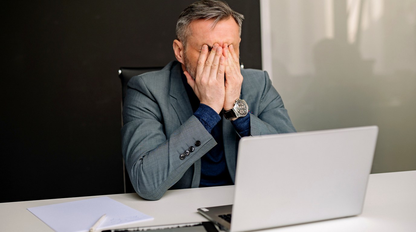 Mann im grauen Sakko sitzt am Schreibtisch und stützt das Gesicht in beide Hände, davor ein aufgeklappter Laptop