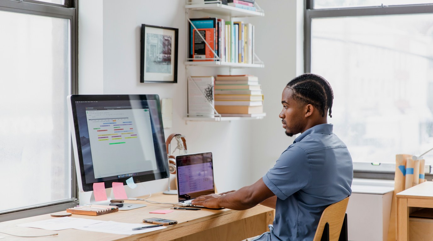 Mann am Schreibtisch arbeitet an einem Laptop und blickt auf einen zweiten Bildschirm mit Software-Oberfläche