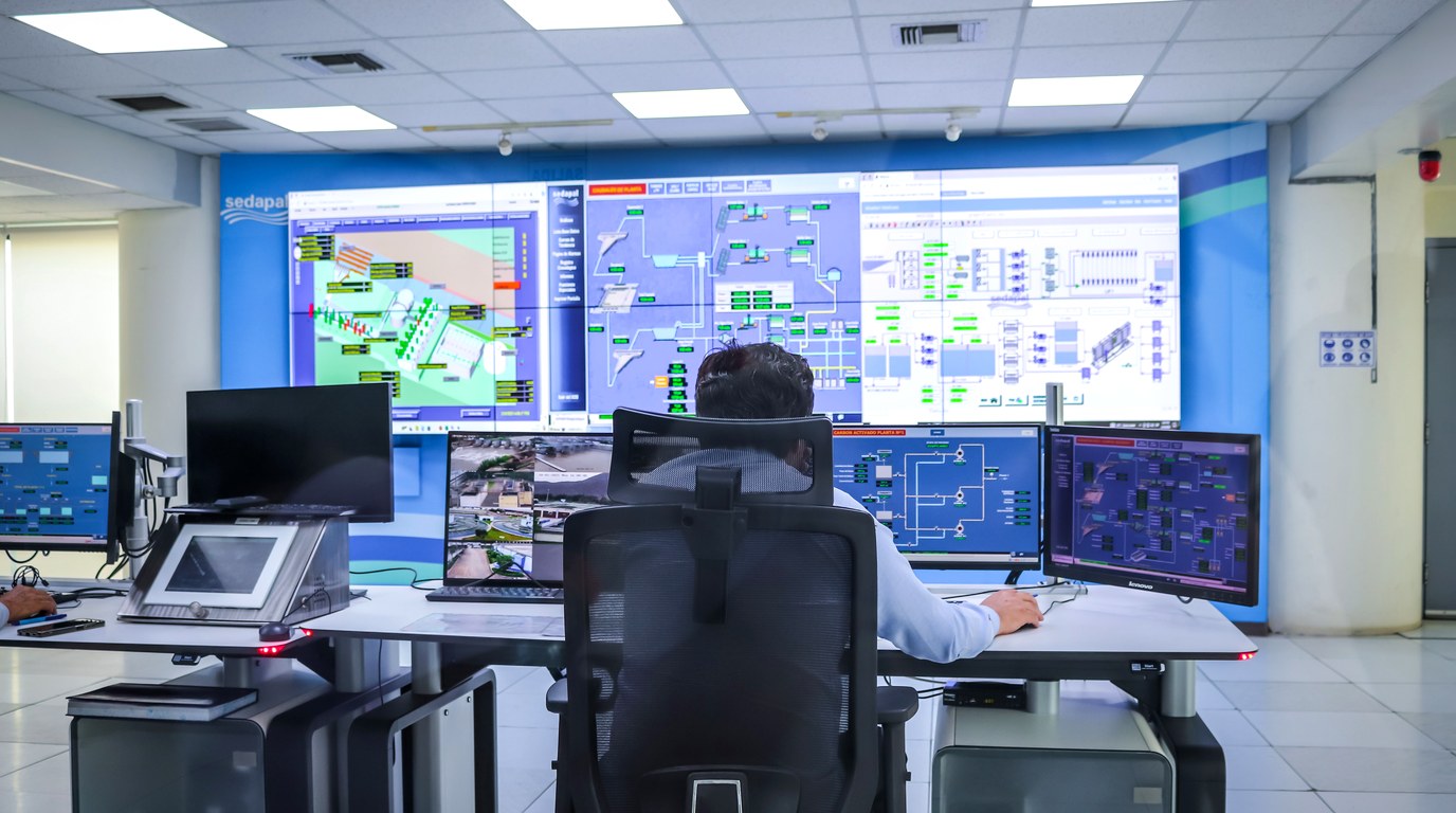 Ein Mensch sitzt allein in einem Leitstand vor einer Monitorwand mit Anlagenschemata und Statusanzeigen