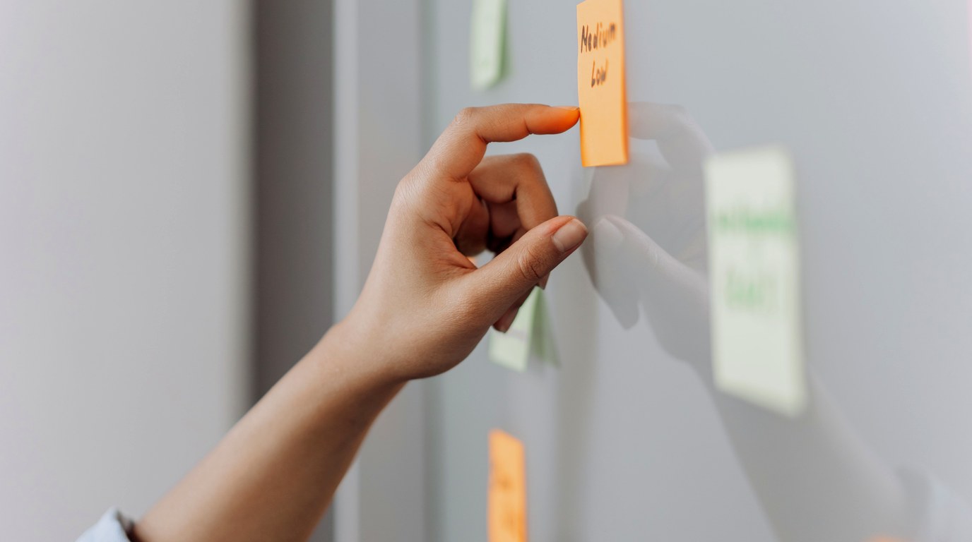 Hand heftet eine orange Haftnotiz an eine Wand, daneben weitere Notizzettel