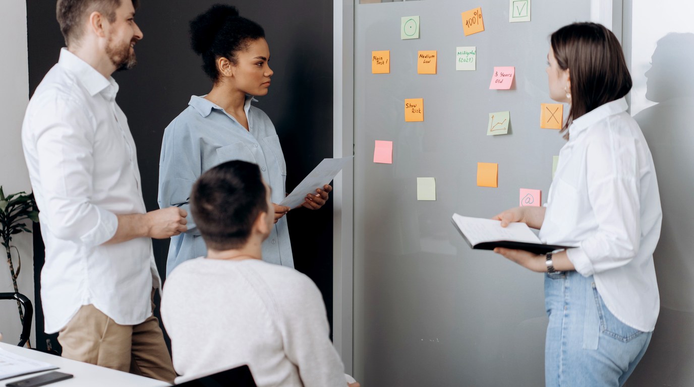 Vier Beschäftigte besprechen in einem Büro Haftnotizen mit Häkchen und Kreuzen an einer Wand