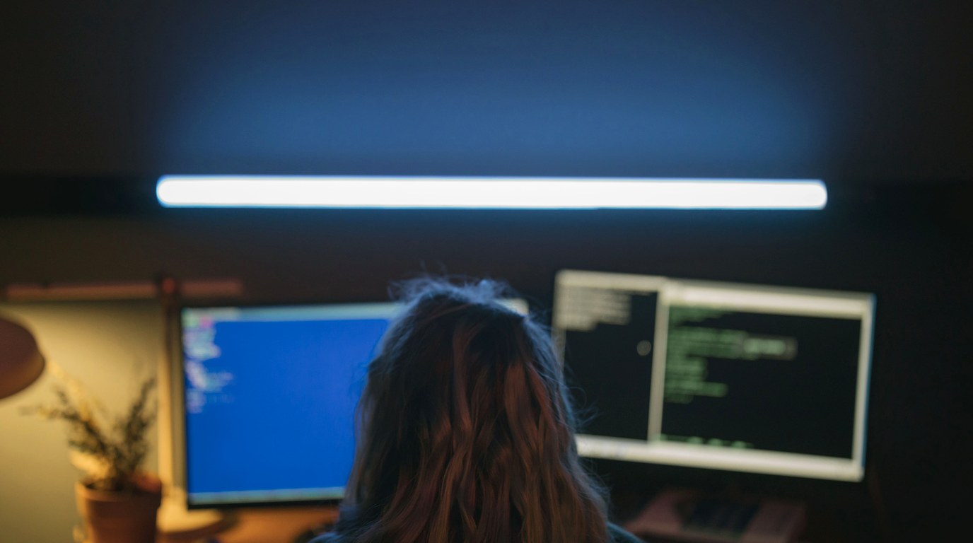 Entwicklerin von hinten vor zwei Monitoren mit Programmcode in einem abgedunkelten Arbeitsraum