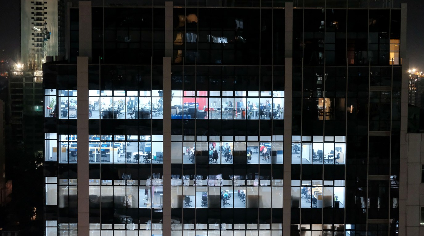 Bürohochhaus bei Nacht, in mehreren beleuchteten Etagen sitzen Menschen an Schreibtischen