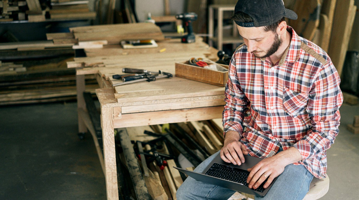 Mann in kariertem Hemd tippt in einer Holzwerkstatt auf einem Laptop