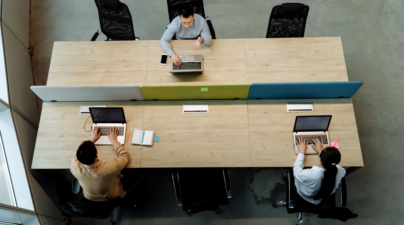 Drei Personen arbeiten an einem langen Bürotisch jeweils an einem eigenen Laptop, von oben fotografiert